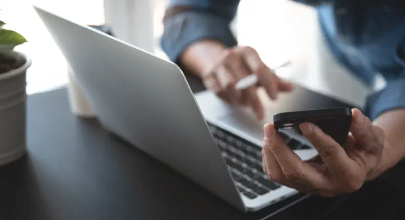 Close-up of a businessman working on a laptop while using a mobile phone, with a digital tablet on the office table